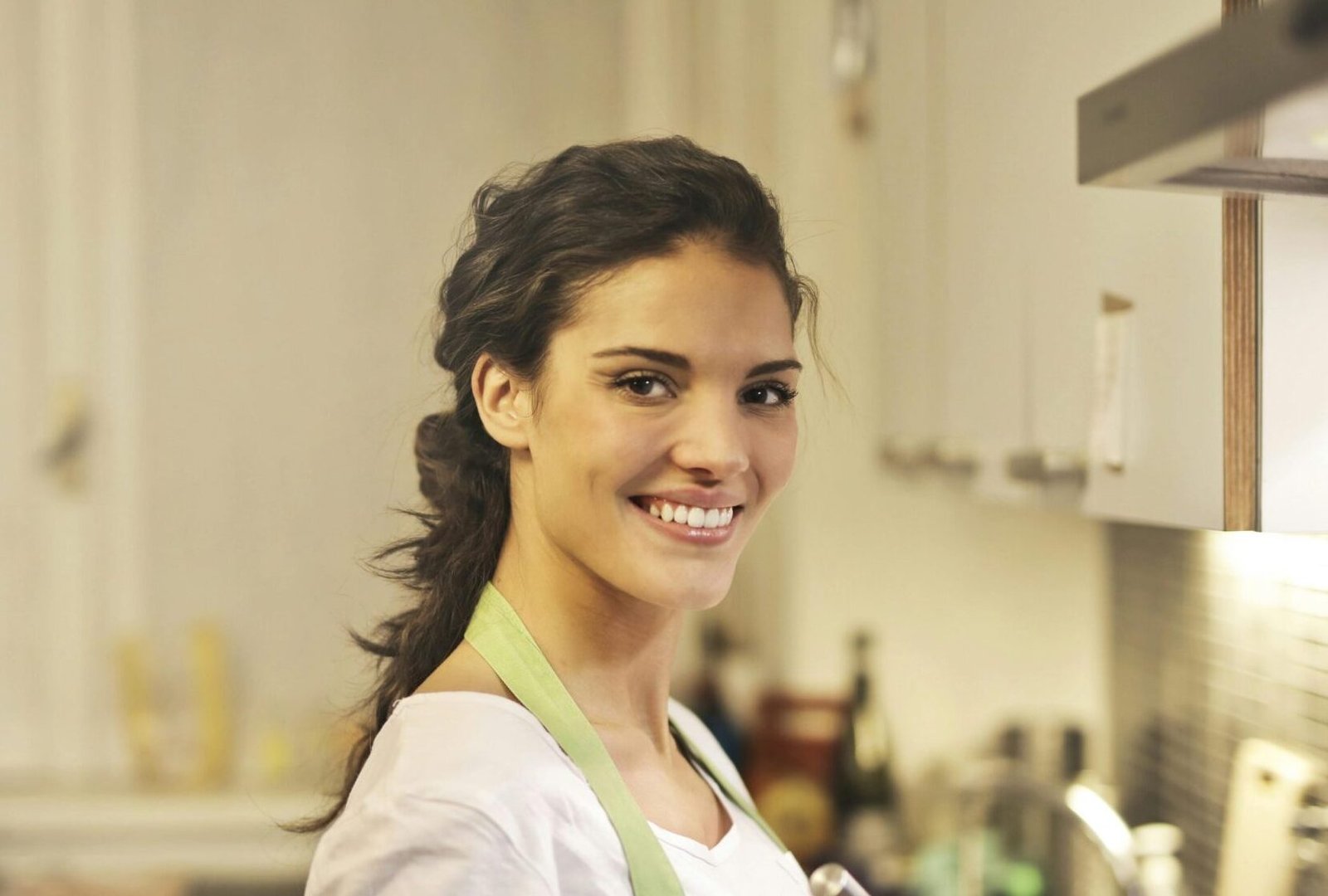 Side view of cheerful female in apron and casual t shirt standing in modern kitchen and mixing ingredients with whisk in stainless bowl while preparing dough