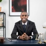 African American businessman smiling in an office environment, seated at a desk.