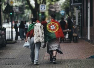 Two people wrapped in a Portuguese flag walking on a city sidewalk.