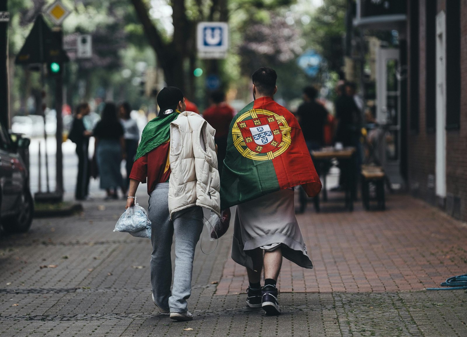 Two people wrapped in a Portuguese flag walking on a city sidewalk.